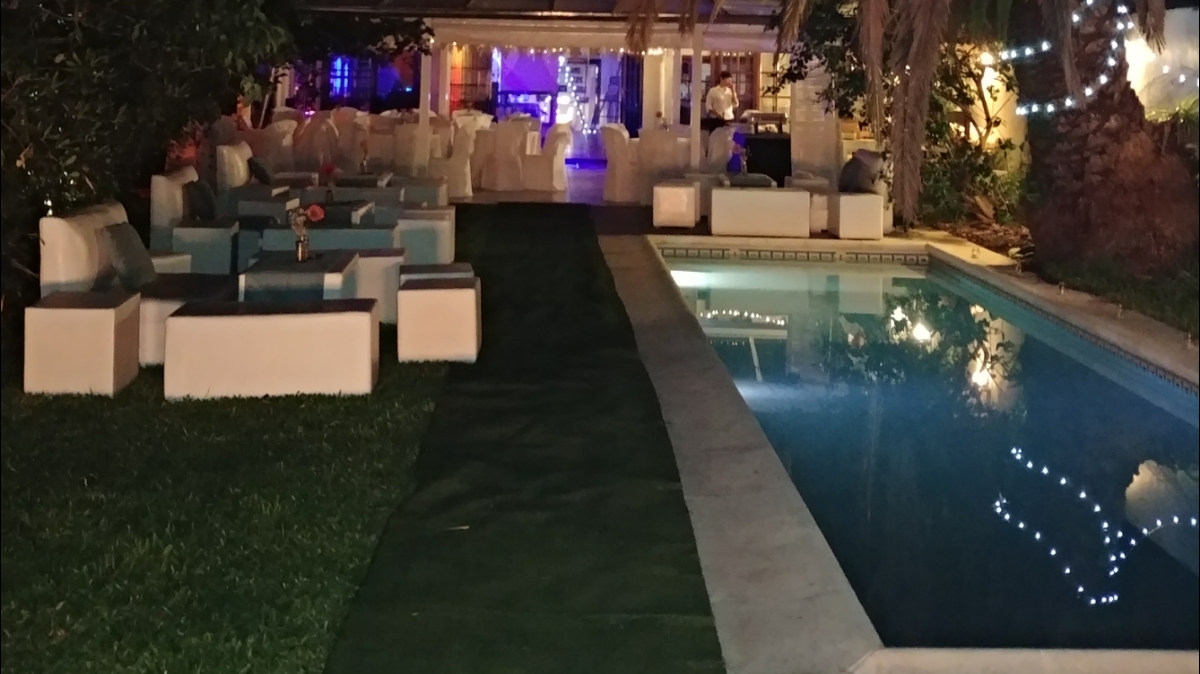 A stunning evening photograph of a luxury private event venue in Buenos Aires Argentina with a lit swimming pool reflecting warm bistro fairy string lights hung above lush green gardens and palm trees, modern outdoor lounge furniture with cocktails on tables, rustic chic wooden quincho pergola warmly lit in the background, festive romantic atmosphere for a wedding or milestone birthday party, shot on 35mm professional architectural photography